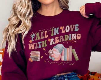 May include: A maroon sweatshirt with the text "Fall in love with reading" in a retro font. A gnome is depicted reading a book, surrounded by books and fall leaves.