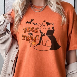 May include: Orange t-shirt with a graphic of a cat wearing a vampire cape and holding a candy cane. The text "Trick or Treat" is printed on the shirt.
