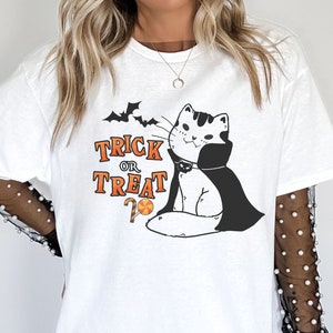 May include: White t-shirt with a black and white illustration of a cat wearing a vampire cape and holding a candy cane. The text 'Trick or Treat' is printed in black with orange accents. The cat is looking to the right and has a mischievous expression.