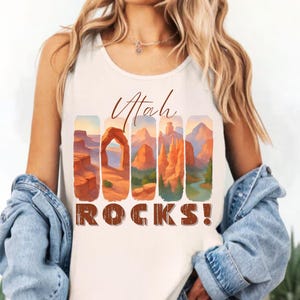 May include: A cream-colored sleeveless tank top featuring a graphic print of Utah landscapes within vertical panels. The text "Utah ROCKS!" is printed in a brown script and block font above and below the colorful desert scenery illustrations.