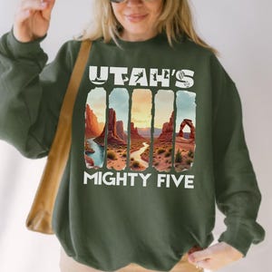 May include: Dark green sweatshirt featuring the text "Utah's Mighty Five" in white. The design includes a landscape illustration of Utah's national parks. The sweatshirt is a comfortable, casual style.