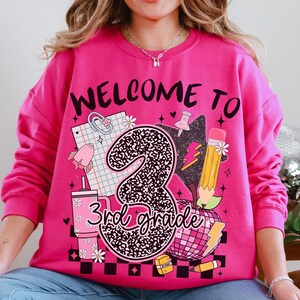 May include: A bright pink sweatshirt with the words "WELCOME TO 3rd grade" in a playful font. The graphic includes a large number 3 with a leopard print design, a pencil, an apple, and other school-themed illustrations.