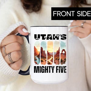 May include: White ceramic mug with a black rim and handle, featuring the text "UTAH'S MIGHTY FIVE" above a colorful landscape design. The design includes images of canyons, rivers, and arches.
