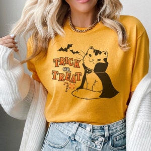 May include: A yellow t-shirt with a black and white illustration of a cat wearing a vampire cape and a bow tie. The text "Trick or Treat" is printed on the shirt with a candy cane and a lollipop.