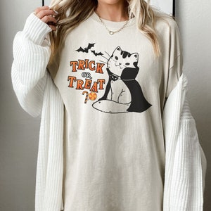 May include: A beige t-shirt with a black and white illustration of a cat wearing a vampire cape. The cat is looking to the right and has a candy cane in its mouth. The text "Trick or Treat" is printed above the cat.