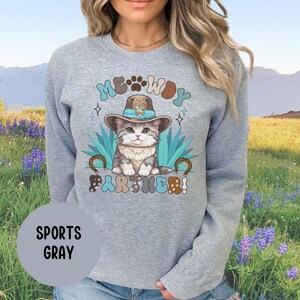 May include: A gray sweatshirt with a cartoon cat wearing a cowboy hat and the text "MEOWDY PARTNER!" printed on the front. The cat is sitting on a cactus with a horseshoe on each side. The sweatshirt is a sports gray color.