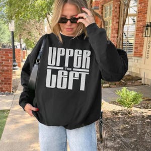 May include: Black sweatshirt with the text "UPPER THE LEFT" in white block letters. The sweatshirt is worn by a person wearing sunglasses and light blue jeans. A black purse is hanging from the person's shoulder.