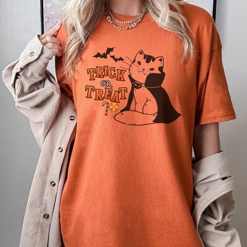 May include: Orange t-shirt with a graphic of a cat wearing a vampire cape and holding a candy cane. The text "Trick or Treat" is printed on the shirt.