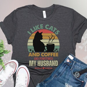 May include: A dark gray t-shirt with a vintage-style graphic that says "I like cats and coffee and sometimes my husband". The graphic features a black cat sitting next to a cup of coffee with steam rising from it. The background is a sunset with orange, yellow, and red colors.