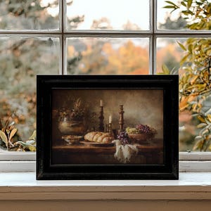 May include: A framed still life painting featuring a table set with bread, grapes, and candles. The painting has a dark wooden frame and is set against a blurred background of fall foliage. The color palette is warm, with browns and golds.