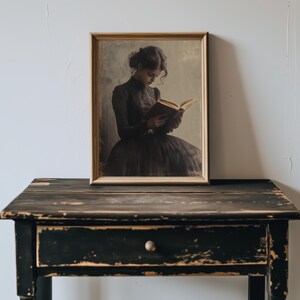 May include: A framed portrait of a person in a dark dress reading a book. The portrait is hanging on a white wall above a black wooden table with a drawer.