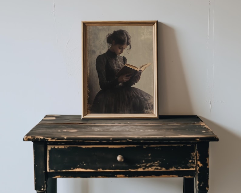 May include: A framed portrait of a person in a dark dress reading a book. The portrait is hanging on a white wall above a black wooden table with a drawer.