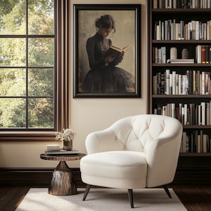 May include: A white tufted armchair with a wooden frame and black legs sits in a room with a window, a bookshelf, and a side table with a vase of flowers. A framed picture of a woman reading a book hangs on the wall.