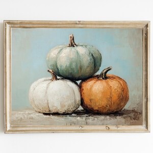 May include: A framed painting of three pumpkins. The top pumpkin is a light blue-green color, the middle pumpkin is white, and the bottom pumpkin is orange. The pumpkins are arranged in a pyramid shape.
