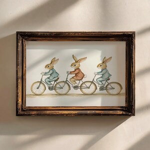 May include: A framed watercolor print features three rabbits riding bicycles. The rabbits wear jackets in blue and brown hues. The artwork is set in a dark brown, ornate frame, adding a touch of vintage charm to the scene.