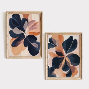 May include: Two framed abstract art prints. Each print features a floral design with navy blue, peach, and blush-colored brushstrokes. The frames are a light wood color, and the art is displayed against a white background.