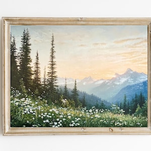 May include: A framed landscape painting depicting a mountain range with a forest in the foreground. The sky is a soft blue with a hint of pink and orange, suggesting a sunset. The mountains are covered in snow and the forest is lush and green. The painting is framed in a light wood frame.