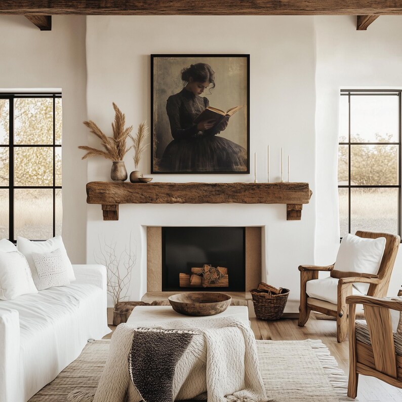 May include: A white living room with a fireplace, a white couch, and two chairs. A framed painting of a woman reading a book hangs above the fireplace. The fireplace has a wooden mantel and a black firebox. The couch has a white throw pillow with a woven design. The chairs have white cushions. The room has a wooden floor and a rug.