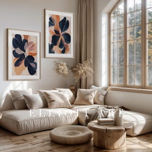 May include: Two framed abstract art prints with botanical designs in navy, peach, and beige tones hang above a beige sectional sofa. The room features a wooden coffee table, a round ottoman, and a large window with natural light.