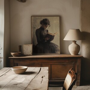 May include: A vintage-style framed print of a woman in a black dress reading a book. The print is hanging on a wall above a wooden dresser with a lamp on the right side.