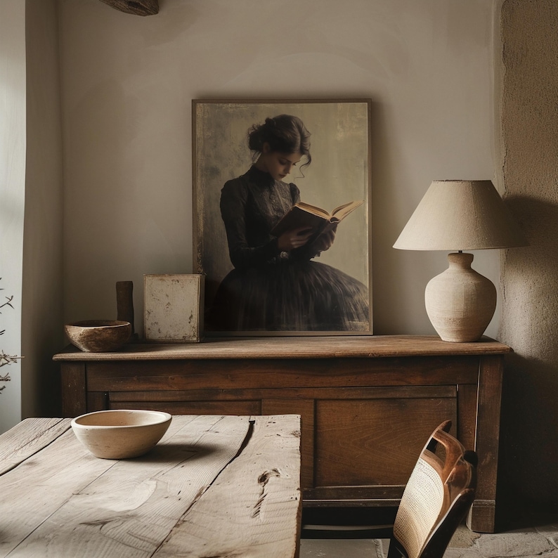May include: A vintage-style framed print of a woman in a black dress reading a book. The print is hanging on a wall above a wooden dresser with a lamp on the right side.