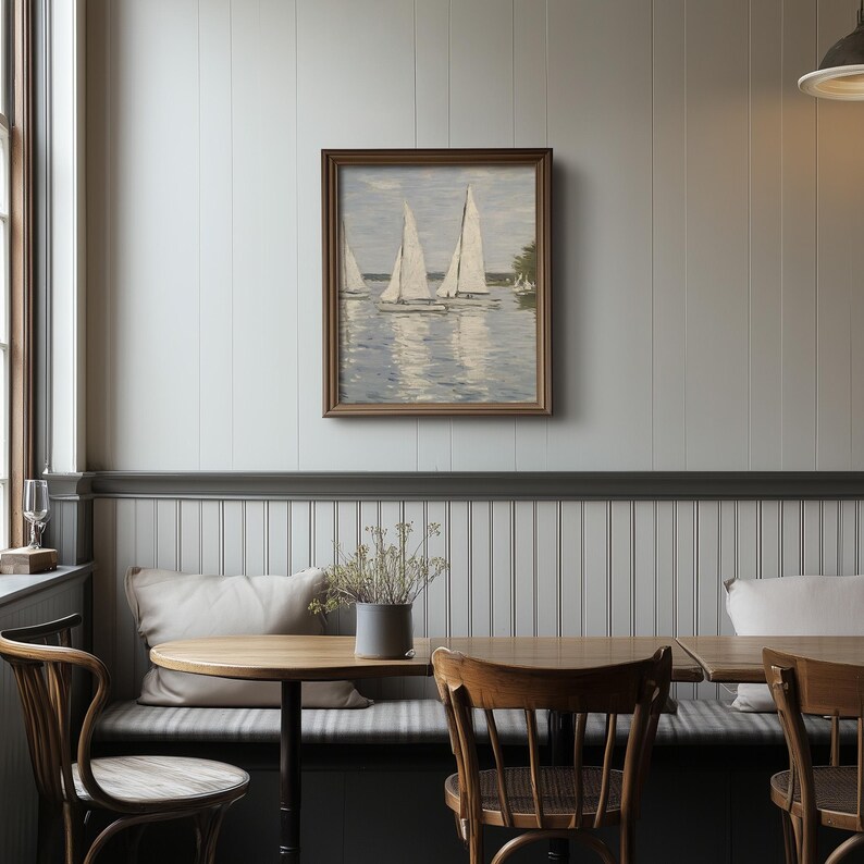 May include: A framed painting depicts sailboats on a body of water. The artwork is in a wooden frame and hangs on a light gray wall. The scene includes white sails and a blue-gray water surface. The painting is displayed in a dining area.