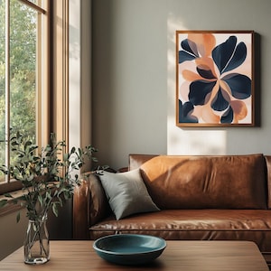 May include: A framed abstract floral art print with navy blue and peach-colored petal shapes. The artwork hangs above a brown leather sofa. A wooden coffee table with a teal bowl and a vase of greenery is in front of the sofa. The text "ELLIOTT ART" is at the bottom.