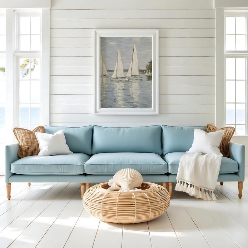 May include: A light blue sofa with a white throw blanket and pillows. A woven coffee table holds a seashell and is in front of the sofa. A framed painting of sailboats hangs above the sofa.