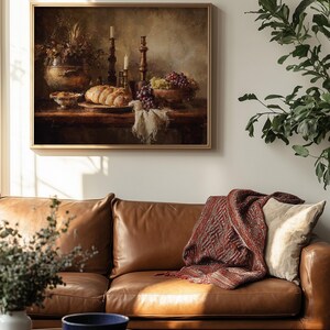 May include: A framed still life painting depicting a table laden with bread, grapes, and candles. The painting hangs above a brown leather sofa with a patterned throw blanket and a cream-colored pillow. A blue bowl sits on a coffee table.