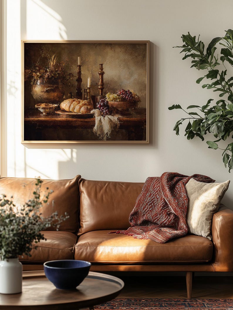 May include: A framed still life painting depicting a table laden with bread, grapes, and candles. The painting hangs above a brown leather sofa with a patterned throw blanket and a cream-colored pillow. A blue bowl sits on a coffee table.