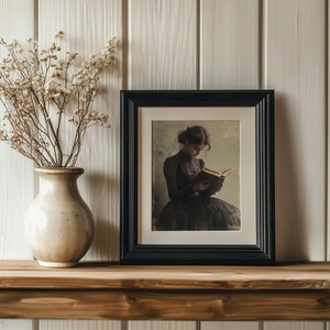 May include: A black framed picture with a woman in a dark dress reading a book. The picture is on a wooden shelf with a vase of dried flowers.