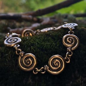 Puede incluir: Pulsera plateada con dijes en forma de espiral. La pulsera descansa sobre musgo verde, con un fondo borroso de árboles y ramas. El diseño evoca un estilo celta o antiguo, con detalles intrincados.