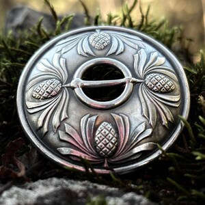 Könnte beinhalten: Silberfarbene, runde Brosche mit aufwendigem floralem Design, das stilisierte Tannenzapfen und Blätter zeigt. Die Brosche hat eine zentrale Öffnung und einen Nadelverschluss. Die detaillierte Metallarbeit ist vor dem Hintergrund von grünem Moos und Stein platziert.