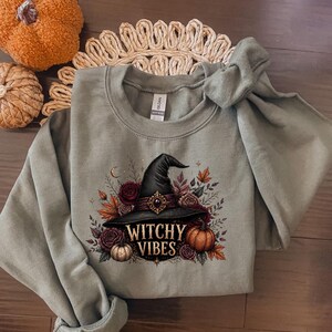 Peut inclure: Un sweat-shirt à col rond vert sauge avec une impression graphique représentant un chapeau de sorcière noir entouré de roses rouge foncé, de feuilles d'automne et de petites citrouilles. Le texte "WITCHY VIBES" est imprimé en lettres dorées.