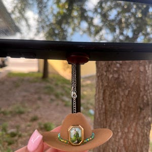 May include: A brown cowboy hat with a green and gold beaded trim hangs from a rearview mirror. A small picture of a saint is in the center of the hat.
