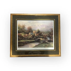 Könnte beinhalten: Gerahmter Kunstdruck einer ruhigen Dorfszene mit einer Steinbrücke über einem Fluss. Das Kunstwerk hat eine warme Farbpalette mit einem goldenen Rahmen und einem dunkelgrünen Passepartout. Der Druck zeigt Häuser, Bäume und Straßenlaternen.