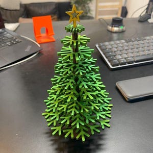 以下が含まれることがあります： 小さな木製のクリスマスツリー。てっぺんに星が付いています。ツリーは緑色に塗られており、黄色い星が付いています。ツリーの高さは約15cmで、黒い表面に置かれています。