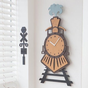Puede incluir: Un reloj de madera con forma de tren con un acabado marrón y negro. La esfera del reloj es de color marrón claro con números y manecillas negros. El reloj está montado sobre una vía de metal negra con una locomotora de madera negra en la parte superior. El reloj tiene una columna de humo azul y blanca sobre el motor. La esfera del reloj dice "STOP ON RED LIGHT" y "RAIL CROSSING ROAD".