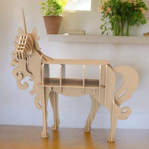 Puede incluir: Una estantería de madera con forma de unicornio con un acabado marrón claro. La estantería tiene cinco estantes y tiene forma de unicornio con un cuerno, una melena y una cola.
