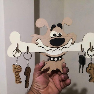 May include: A wooden key holder shaped like a cartoon dog with a bone in its mouth. The dog is brown and has a black nose and eyes. The bone is white and has two key rings on each side. The key holder is hanging on a wall.