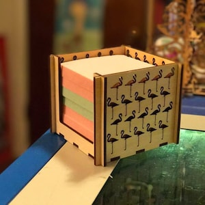 Può includere: Un porta note in legno con un motivo a forma di fenicottero. Il porta note contiene un pacco di note adesive colorate.