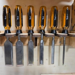 Könnte beinhalten: Ein Wandorganizer aus Holz mit sieben Steckplätzen zur Aufbewahrung von Meißeln. Der Organizer ist aus hellem Holz mit dunkelbraunen Akzenten. Die Meißel sind alle aus Metall mit gelben und schwarzen Griffen.