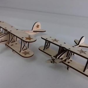Puede incluir: Dos aviones de madera modelo con detalles marrones y estrellas. Los aviones están hechos de madera clara y tienen un diseño simple.