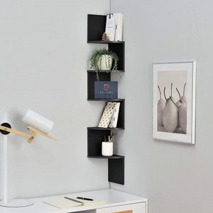 Puede incluir: Estantería de esquina negra con cinco estantes. Los estantes están escalonados y en ángulo. El estante superior tiene algunos libros y una planta pequeña. El segundo estante tiene una planta en maceta. El tercer estante tiene un pequeño letrero con el texto "DecorDen". El cuarto estante tiene una pila de libros. El quinto estante tiene un pequeño jarrón con lápices. La estantería está montada en una pared blanca.