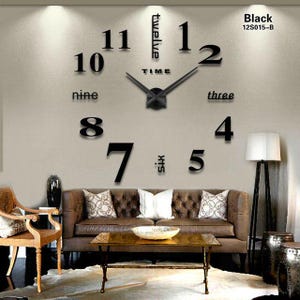 May include: A large black 3D clock with removable numbers and hands. The clock is mounted on a white wall with a brown leather sofa and a lamp in the background. The clock numbers are 1, 2, 3, 4, 5, 6, 7, 8, 9, 10, 11, and 12. The clock also has the words "twelve" and "time" on it.