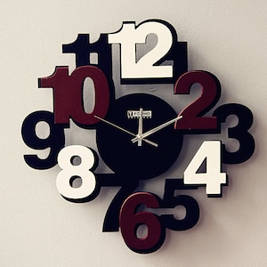 May include: A black and white clock with large, colorful numbers. The numbers are arranged in a circle around a smaller black clock face with silver hands. The clock face has the text "TFCIND" printed on it.