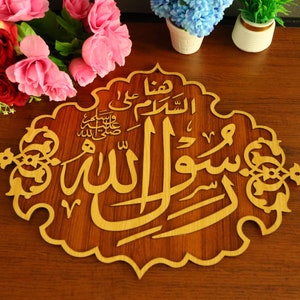 May include: A wooden wall decoration with intricate carvings in a gold colour. The Arabic calligraphy reads "Allah" and "Muhammad" in a decorative font. The background is a dark brown wood.