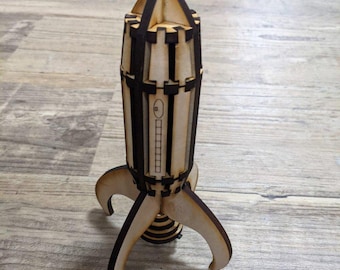Op de afbeelding: Een houten raket speelgoed met een zwart-wit gestreept lichaam en drie poten. De raket heeft een klein raampje en een gedetailleerd ontwerp.