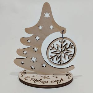 Peut inclure: Une décoration de sapin de Noël en bois avec un flocon de neige blanc suspendu. L'arbre est décoré d'étoiles blanches et la base porte l'inscription "С Новым годом!" qui se traduit par "Bonne année!" en français.