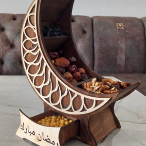 Op de afbeelding: Een houten snackkom in de vorm van een halve maan met drie compartimenten en een lade. De kom is versierd met ingewikkelde uitsnijdingen en de woorden "Ramadan Mubarak" zijn gegraveerd op de lade. De kom is gevuld met gedroogd fruit en noten.